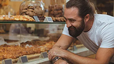 Eine Person lehnt an einer Glasvitrine in einer Bäckerei und betrachtet eine Auswahl an Gebäck und Backwaren, darunter Croissants und Kekse.