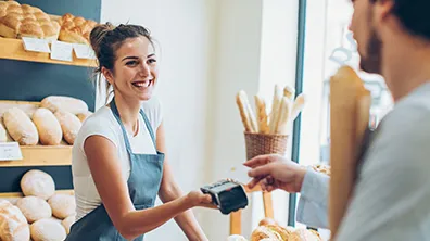 Kunde tätigt eine Kartenzahlung bei einer Bäckereimitarbeiterin, die ein Zahlungsterminal hält, mit Brotauslagen im Hintergrund.