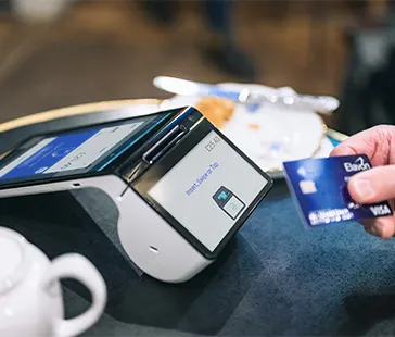 Nahaufnahme einer Person, die eine Kreditkarte in der Nähe eines modernen Zahlungsterminals auf einem Café-Tisch mit Teekanne und Teller hält.