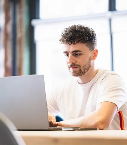 Person arbeitet an einem Laptop an einem Schreibtisch in heller, moderner Umgebung mit großen Fenstern im Hintergrund.