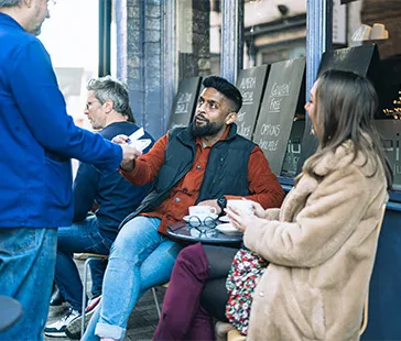 Personen sitzen vor einem Café, während eine Person eine Kartenzahlung an eine andere am kleinen Tisch vornimmt.