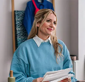 Person trägt einen hellblauen Pullover und hält Stoffmuster in einem kreativen Arbeitsbereich mit Regalen und hängender Kleidung im Hintergrund.