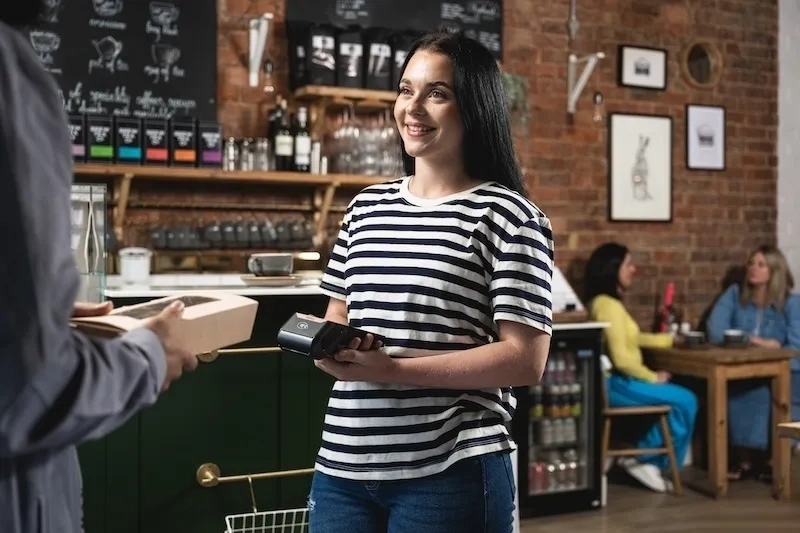 Eine Person in einem gestreiften Hemd hält ein Zahlungsterminal in einem Café während einer Transaktion mit Regalen, Kreidetafeln und mit Menschen, die am Tisch plaudern, im Hintergrund